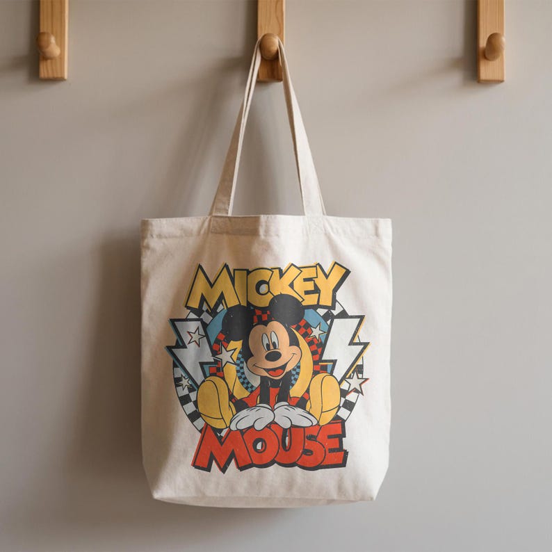 May include: A cream-colored tote bag featuring a colorful graphic of Mickey Mouse. The design includes the words "Mickey Mouse" in a retro font, with lightning bolts and stars. The bag has long handles for carrying.