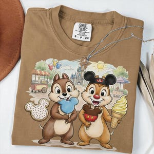 May include: Brown Comfort Colors t-shirt featuring Chip and Dale with Disney treats and a castle backdrop. The shirt includes a necklace and a small tag. The graphic includes a Mickey Mouse ice cream bar, a blue popsicle, and a cookie.