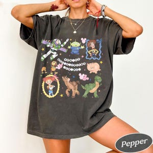 Pu&ograve; includere: T-shirt grigio scuro con personaggi di cartoni animati colorati di un popolare film d'animazione. La maglietta include immagini di un ranger spaziale, un alieno, un cowboy, un maiale, un dinosauro, un cavallo e una ragazza. Il testo "You've Got a Friend in Me" &egrave; anche sulla maglietta.