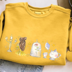 以下が含まれることがあります： マスタードイエローのスウェットシャツ。おとぎ話がテーマのイラストが描かれています。デザインには、燭台、茶色の髪のキャラクター、ガラスドームに入ったバラ、本、ティーカップ、ティーポットが含まれています。デザインは黒で縁取られています。