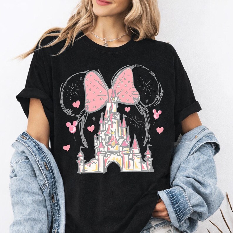 May include: A black short-sleeved t-shirt featuring a graphic print of a castle with a large pink polka-dot bow above it. The design includes small pink hearts, mouse-ear shaped balloons, and firework outlines in a sketch-like artistic style.