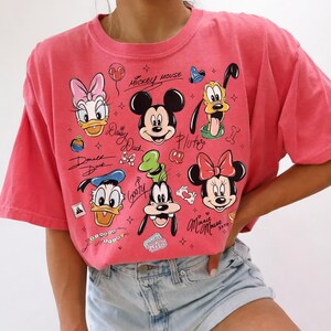 May include: A coral-colored t-shirt featuring a graphic of Mickey Mouse, Minnie Mouse, Donald Duck, Daisy Duck, Goofy, and Pluto. The design includes character names and other Disney-themed elements.