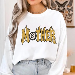 May include: A white ceramic mug with a black handle and the text "But First, Coffee" in a bold, black font. The mug is set against a blurred background, suggesting a cozy setting.