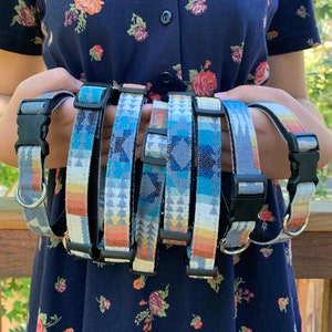 May include: Several dog collars with a Southwestern-inspired pattern in shades of blue, gray, orange, and yellow are displayed. Each collar has a black plastic buckle and a silver metal D-ring for leash attachment. The collars are held up against a blurred background.