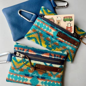 May include: Two small zippered pouches with a southwestern pattern and a leather tag that says "RBO." The pouches have a blue and white zipper and a brown leather strap with a carabiner clip. One pouch has a sticker inside with a coffee cup, a van, and a backpack.