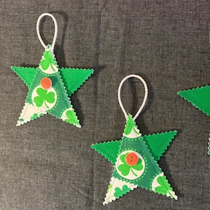 Puede incluir: Tres adornos verdes en forma de estrella con lazos blancos para colgar. Cada estrella tiene un borde verde festoneado y un centro estampado con tréboles y un pequeño botón naranja. Los adornos son probablemente para el Día de San Patricio.