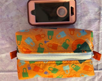 May include: An orange pencil case with a white zipper and a pattern of colorful locks and keys. A pink and brown phone case sits above the pencil case on a patterned surface.