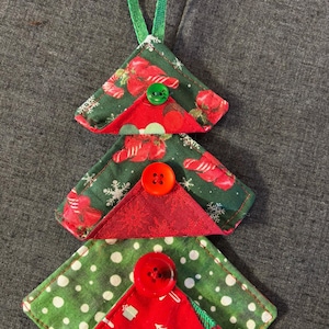 May include: A handmade Christmas tree ornament. The tree is constructed from three layers of fabric in green and red patterns, with a green loop for hanging. Each layer is adorned with a colorful button. The ornament is approximately 5 inches tall.
