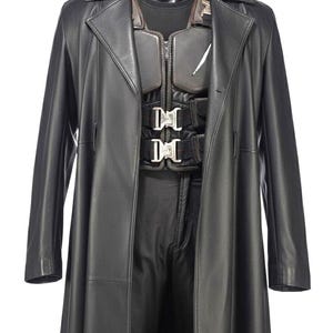 May include: Black leather trench coat with a matching vest and trousers set. The vest has a zip closure and silver buckles. The outfit is displayed on a black mannequin.