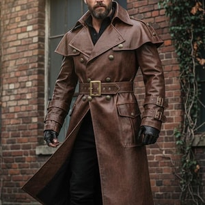 Puede incluir: Un abrigo largo de cuero marrón con un diseño cruzado, un cinturón con hebilla rectangular y charreteras. El abrigo presenta múltiples bolsillos y botones decorativos. Las mangas tienen detalles de hebilla. La persona lleva guantes negros y pantalones negros.