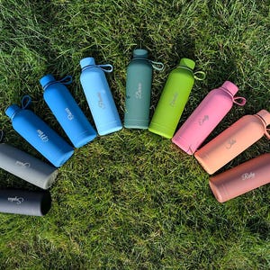 May include: A collection of ten matte-finish water bottles arranged in a semi-circle. Each bottle features a unique colour, including black, grey, blue, green, lime, pink, peach, and coral, with a personalised name engraved in white script.