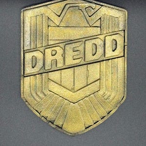 May include: A gold-colored, shield-shaped emblem with the word "DREDD" in bold, capital letters. The emblem has a textured surface and a geometric design, with a dark background.