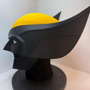 Peut inclure: Un casque de Wolverine noir et jaune, avec un masque noir textur&eacute; et des protub&eacute;rances distinctives en forme d'oreilles. Le dessus du casque est jaune vif. Le casque est expos&eacute; sur un support noir.