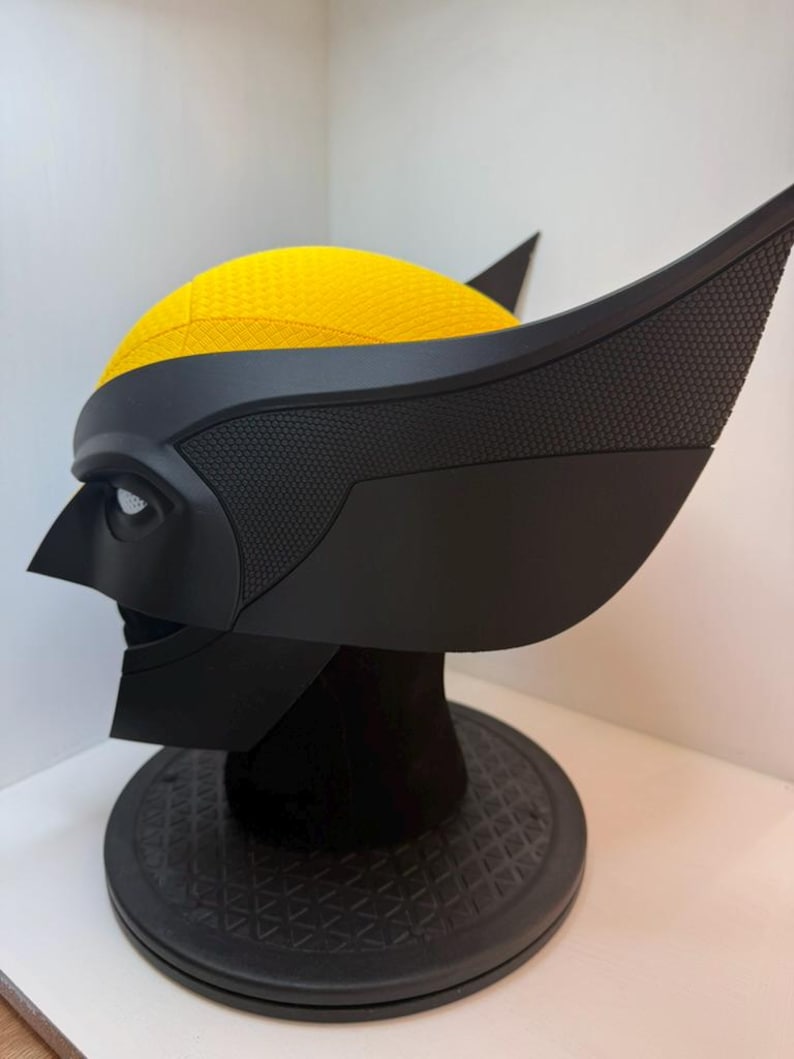 Peut inclure: Un casque de Wolverine noir et jaune, avec un masque noir textur&eacute; et des protub&eacute;rances distinctives en forme d'oreilles. Le dessus du casque est jaune vif. Le casque est expos&eacute; sur un support noir.