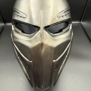 Könnte beinhalten: Ein metallisch silberner Vollvisierhelm mit einem schlanken, kantigen Design. Der Helm verfügt über Augenöffnungen mit dunklem Innenraum, einen markanten Mittelgrat und detaillierte Oberflächenakzente. Die Gesamtästhetik deutet auf ein futuristisches oder taktisches Thema hin.