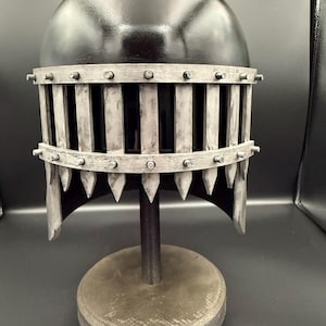 Peut inclure: Un casque noir doté d'une protection faciale argentée en forme de cage avec des barres verticales et des pointes. La protection est fixée par de petits rivets le long des bandes horizontales, créant un aspect structuré et protecteur.