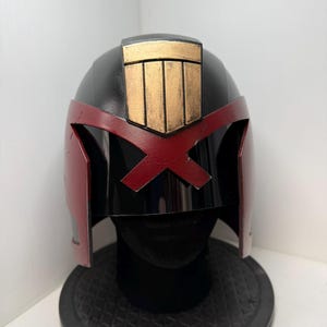 May include: A black and red helmet with a gold emblem on top. The helmet has a red X-shaped design across the visor and red side panels. The helmet is displayed on a black stand.