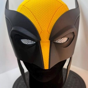 May include: A black and yellow Wolverine mask, with a textured yellow top and black facial features. The mask has pointed ear-like extensions and eye openings with white mesh detailing. The mask is displayed on a black stand.