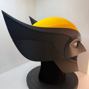Peut inclure: Un masque de Wolverine noir et jaune, avec les oreilles pointues et les traits du visage caract&eacute;ristiques. Le masque est pr&eacute;sent&eacute; sur un socle noir, mettant en valeur les surfaces textur&eacute;es et les &eacute;l&eacute;ments de design.