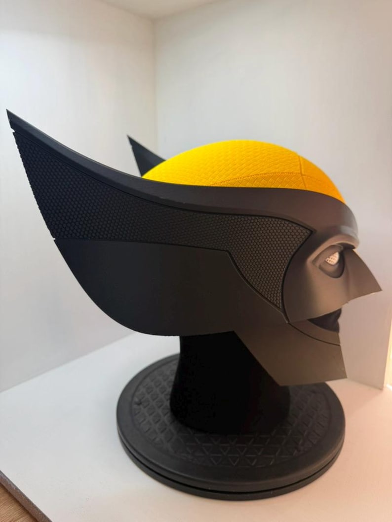 Peut inclure: Un masque de Wolverine noir et jaune, avec les oreilles pointues et les traits du visage caract&eacute;ristiques. Le masque est pr&eacute;sent&eacute; sur un socle noir, mettant en valeur les surfaces textur&eacute;es et les &eacute;l&eacute;ments de design.