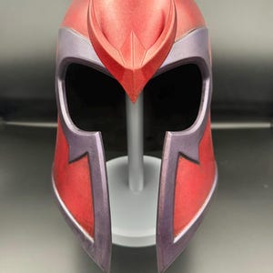 Peut inclure: Un casque intégral avec une finition rouge métallique, doté d'une crête centrale proéminente et d'accents violets autour de l'ouverture pour les yeux et de la protection faciale inférieure. Le casque présente un design lisse et profilé.