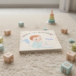 May include: A children's book with the text "Happy 1st Birthday" and the name "Theo" on the cover. The illustration shows a baby with a birthday cake. Wooden blocks and a stacking toy are scattered around the book.