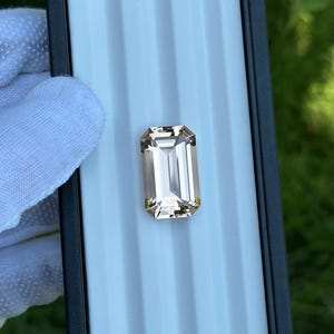 May include: A rectangular, faceted gemstone with a light brown hue. The gem is set against a light blue background, with a black border. The gem is held by a gloved hand. The gem is a beautiful example of fine jewelry.