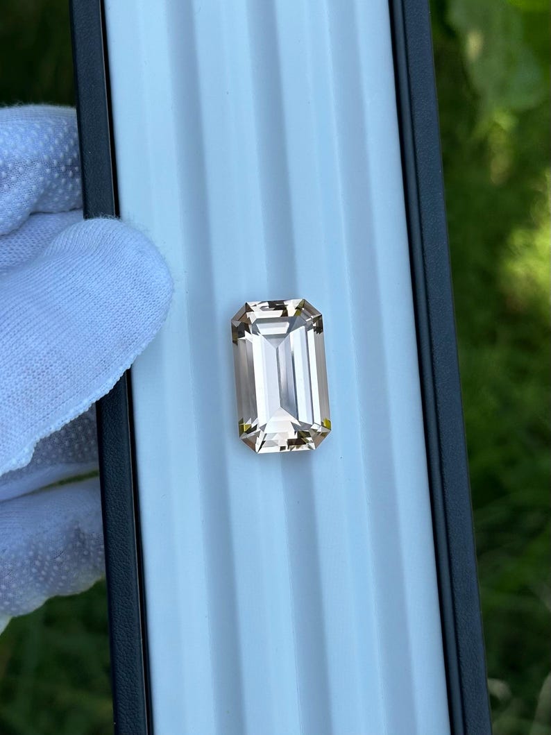 May include: A rectangular, faceted gemstone with a light brown hue. The gem is set against a light blue background, with a black border. The gem is held by a gloved hand. The gem is a beautiful example of fine jewelry.