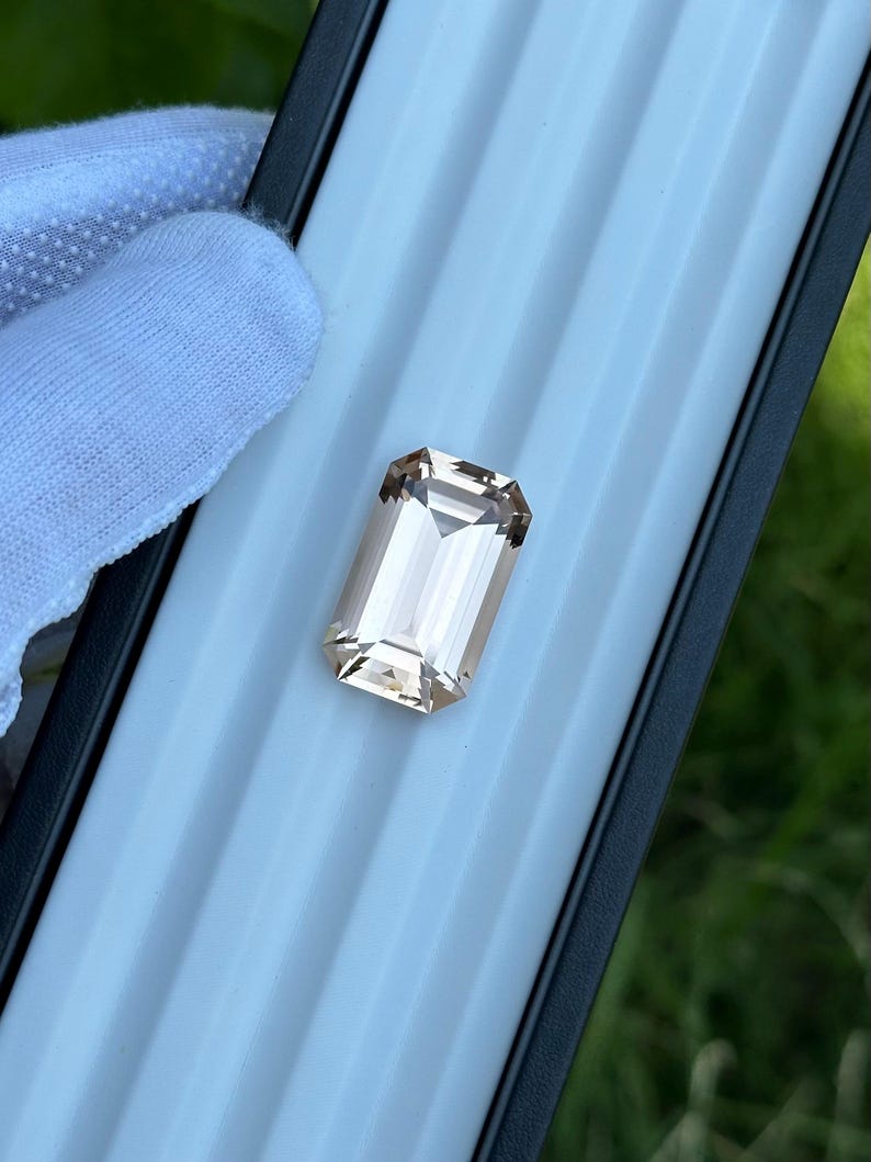 May include: A rectangular, light peach-colored gemstone with an emerald cut. The gem is held in a black and light blue case, with a white-gloved hand visible in the background. The gem has a high degree of clarity and brilliance.