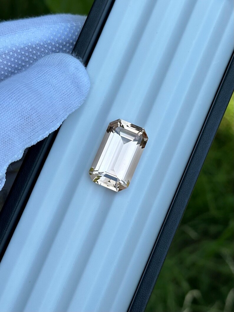 May include: A rectangular, light peach-colored gemstone with a faceted cut. The gem is set in a black case with a light blue interior. The gem is held by a gloved hand.