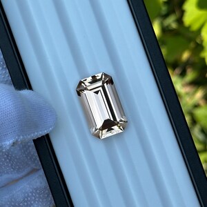 May include: A rectangular, light brown gemstone with an emerald cut. The gem is set against a white background within a black-edged tray. The facets of the stone reflect light, creating a sparkling effect. The overall presentation suggests a focus on jewelry or gemstones.