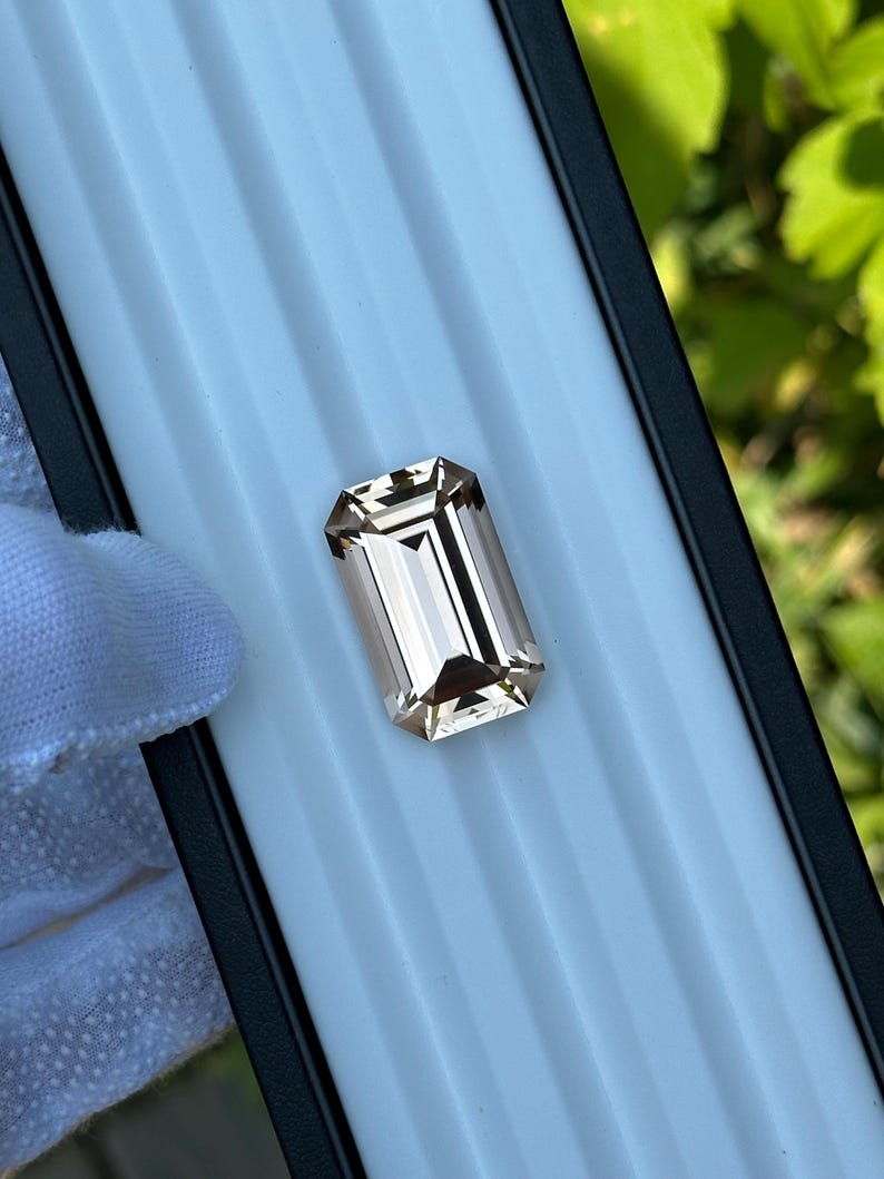 May include: A rectangular, light brown gemstone with an emerald cut. The gem is set against a white background within a black-edged tray. The facets of the stone reflect light, creating a sparkling effect. The overall presentation suggests a focus on jewelry or gemstones.