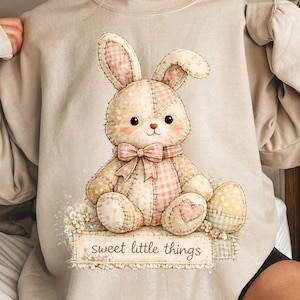May include: Beige sweatshirt featuring a stitched bunny design with a pink bow and a matching checkered scarf. The bunny sits on a floral base with the text "sweet little things". The design includes a pastel Easter egg.