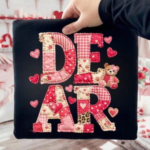 May include: Black square pillow with the word "DEAR" appliqued in patchwork fabric. The letters are red, pink, and cream with heart and floral patterns. A small teddy bear and heart accents complete the design.