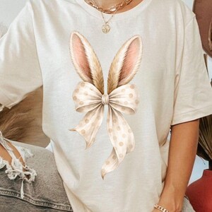 May include: Cream-colored t-shirt featuring a watercolor design of bunny ears and a bow. The bunny ears are light brown with pink inner ears, and the bow is cream-colored with small, light brown polka dots. The shirt is a casual, everyday wear item.