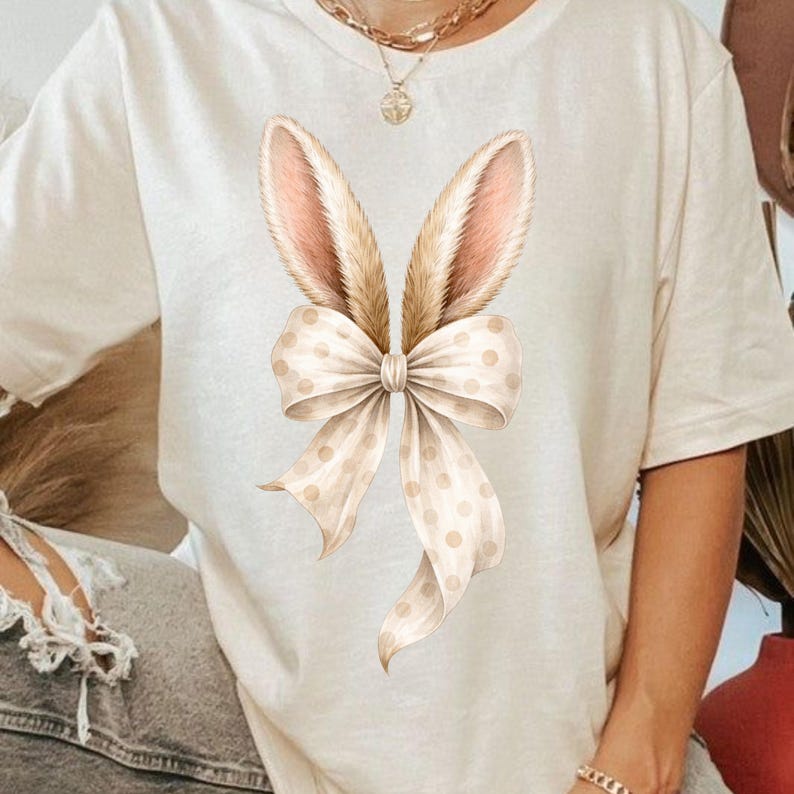May include: Cream-colored t-shirt featuring a watercolor design of bunny ears and a bow. The bunny ears are light brown with pink inner ears, and the bow is cream-colored with small, light brown polka dots. The shirt is a casual, everyday wear item.