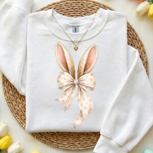 May include: White sweatshirt featuring a bunny ears and bow design. The bunny ears are tan with pink inner ears, and the bow is cream-colored with tan polka dots. A gold necklace with a heart pendant is also visible.