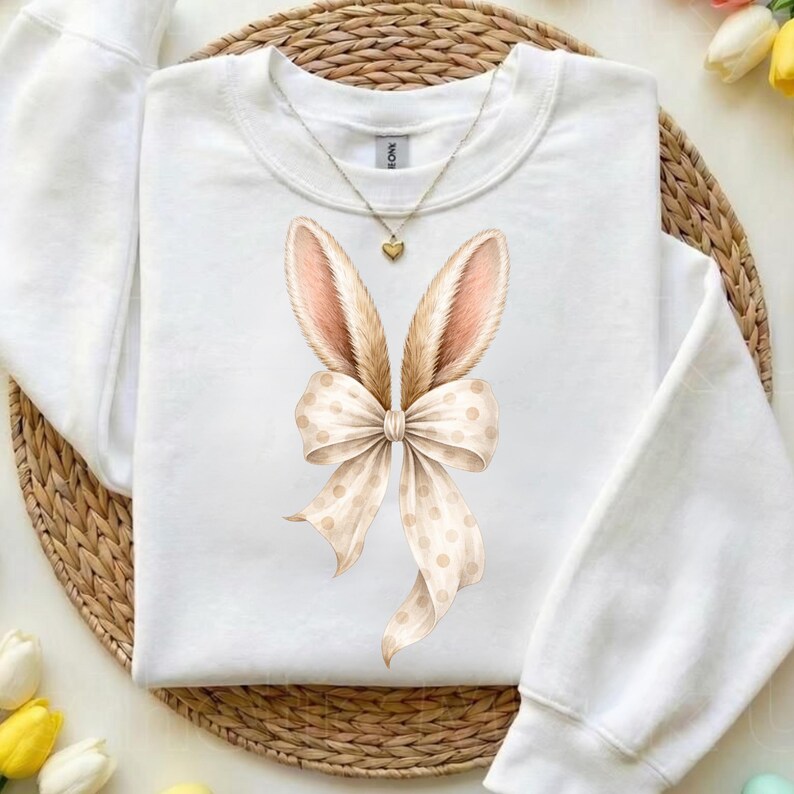 May include: White sweatshirt featuring a bunny ears and bow design. The bunny ears are tan with pink inner ears, and the bow is cream-colored with tan polka dots. A gold necklace with a heart pendant is also visible.