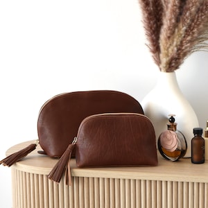 May include: Two brown leather cosmetic bags with gold zippers and tassel pulls sit on a light wood table. A white vase with dried pampas grass, perfume, and skincare products are also on the table.