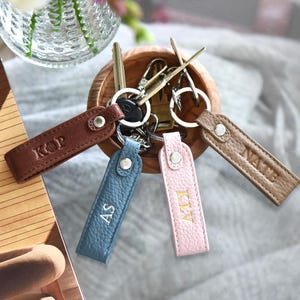 May include: A collection of personalized leather keychains in brown, blue, and pink, each with initials. The keychains are attached to metal key rings and are arranged in a wooden bowl. A book and flowers are in the background.