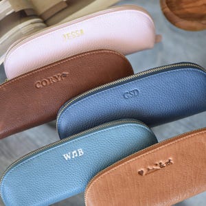 May include: Five semi-circular zippered pouches made of textured leather in pink, brown, and blue shades. Each pouch features a gold-tone zipper and custom embossed lettering or symbols, including JESS, CORY, GSD, WJB, and a heart with an ampersand.