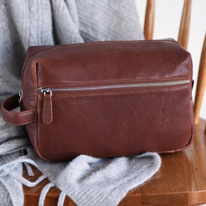 May include: A brown leather toiletry bag with a silver zipper closure and a small handle on the side. The bag is rectangular and sits on a wooden chair with a gray scarf draped over it.