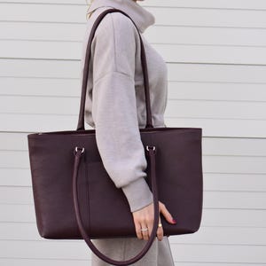 May include: A burgundy leather tote bag with long handles. The bag has a rectangular shape and a zippered top. The woman is wearing a gray sweatshirt and sunglasses.