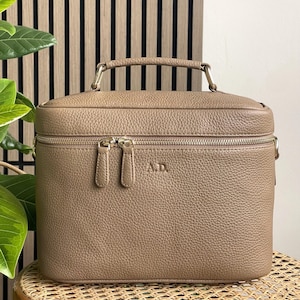 May include: A taupe-colored, rectangular makeup case with a top handle and a wraparound zipper. The case has a textured surface and gold-toned hardware. The initials "A.D." are embossed on the front.