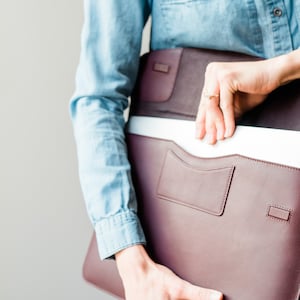 May include: A burgundy leather laptop sleeve with a zipper closure and a front pocket. The sleeve is open and a laptop is partially visible inside.