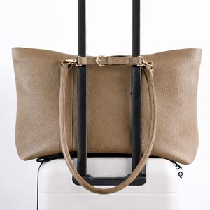 May include: A taupe leather tote bag with a shoulder strap rests on top of a white rolling suitcase. The suitcase has a black handle. The tote bag has gold-colored hardware.