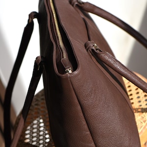 May include: A brown leather tote bag with a gold zipper closure. The bag has two handles and a shoulder strap. The leather has a textured appearance. The bag is sitting on a wooden chair.