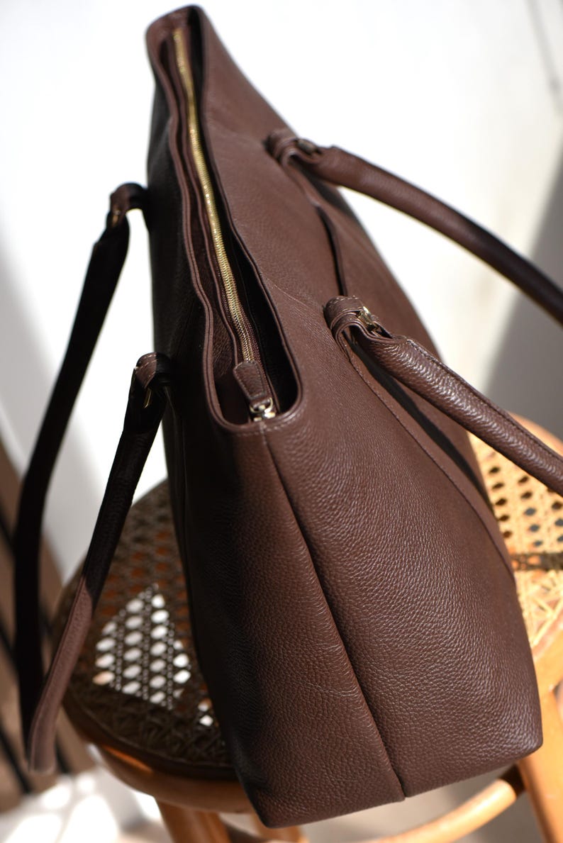 May include: A brown leather tote bag with a gold zipper closure. The bag has two handles and a shoulder strap. The leather has a textured appearance. The bag is sitting on a wooden chair.