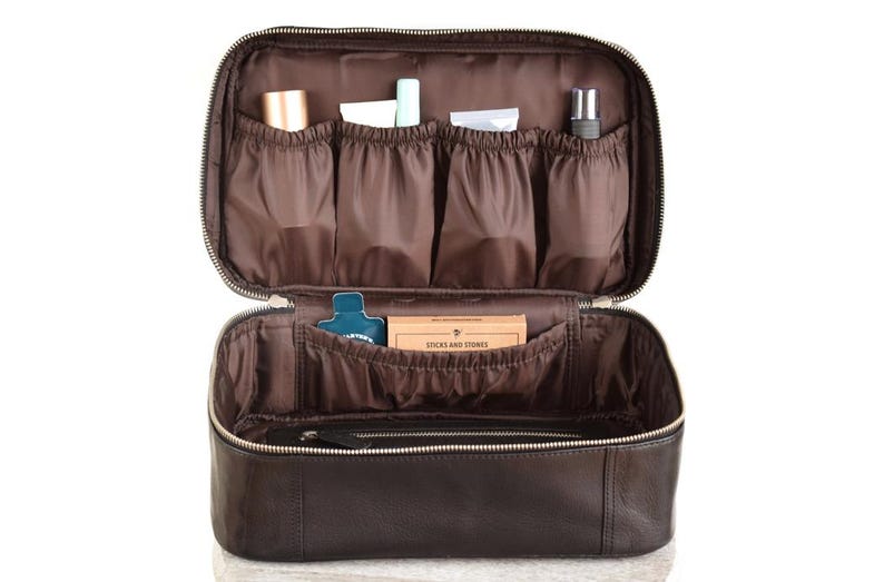 A black full grain leather toiletry bag with a zipper closure. The bag is open and the interior is lined with brown wipe clean fabric. The bag has multiple pockets and compartments for storing toiletries. The bag is sitting on a white surface.