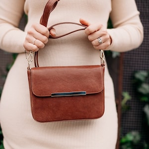 May include: A brown leather shoulder bag with a silver metal clasp. The bag has a long strap and is worn by a person in a white dress.